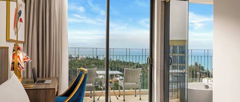 A hotel room with a large sliding glass door to a balcony, a desk with a laptop, a blue chair, and a bed.