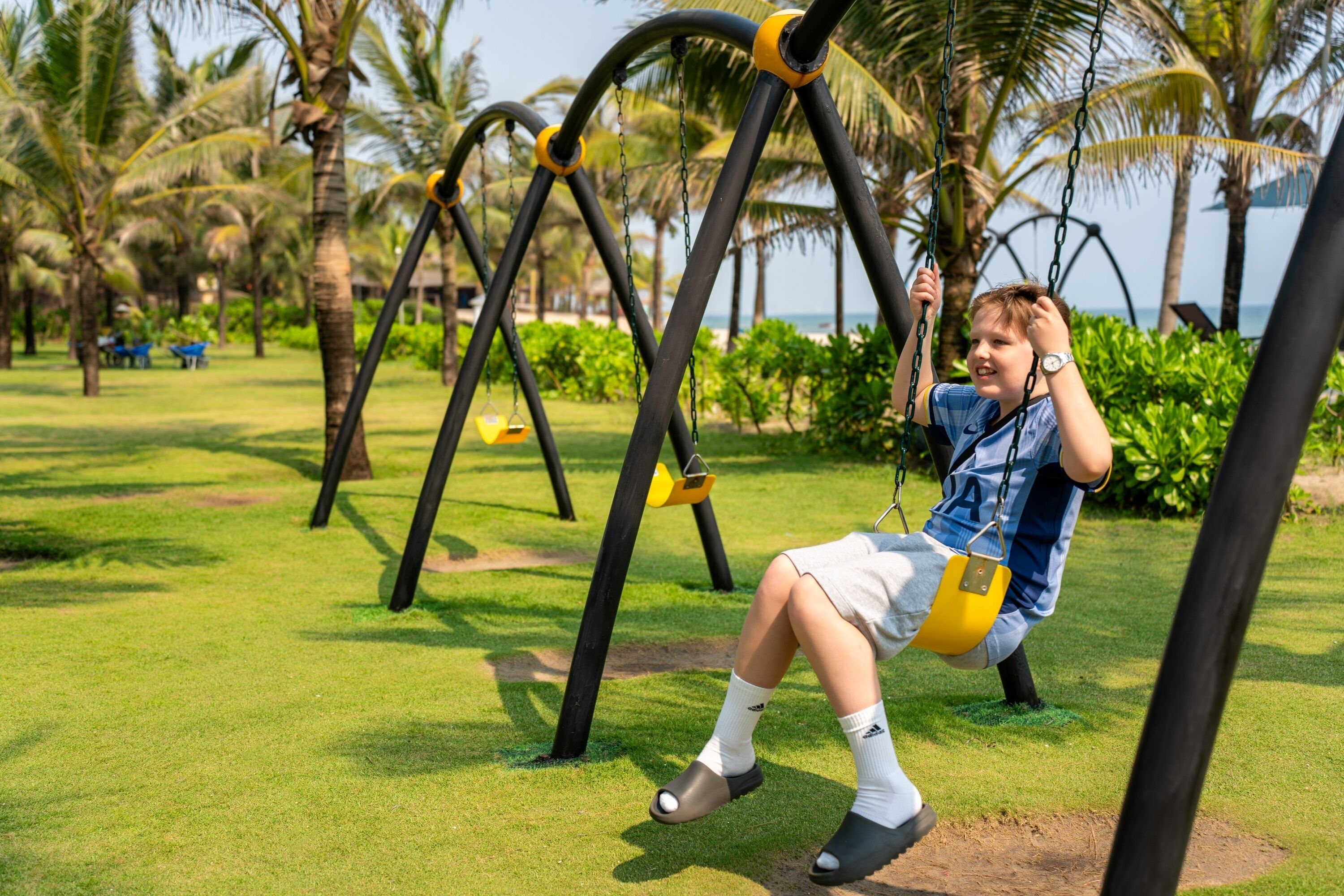 Ein Kind schwingt sich in einem Park mit Palmen und Blick auf das Meer.