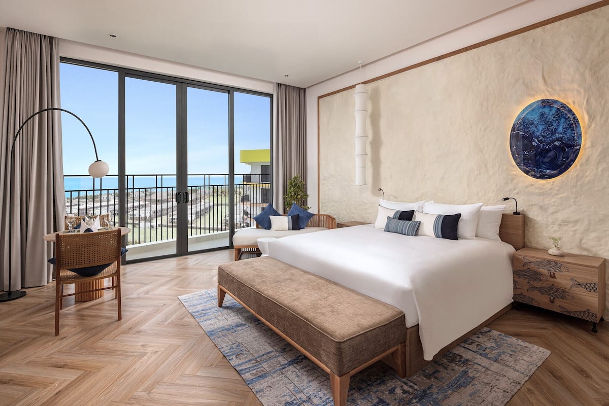 A modern bedroom with a large bed, a bench, a desk, and a view of the sea through large windows.