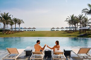 A couple sitting by an infinity pool with a view of the beach and palm trees.