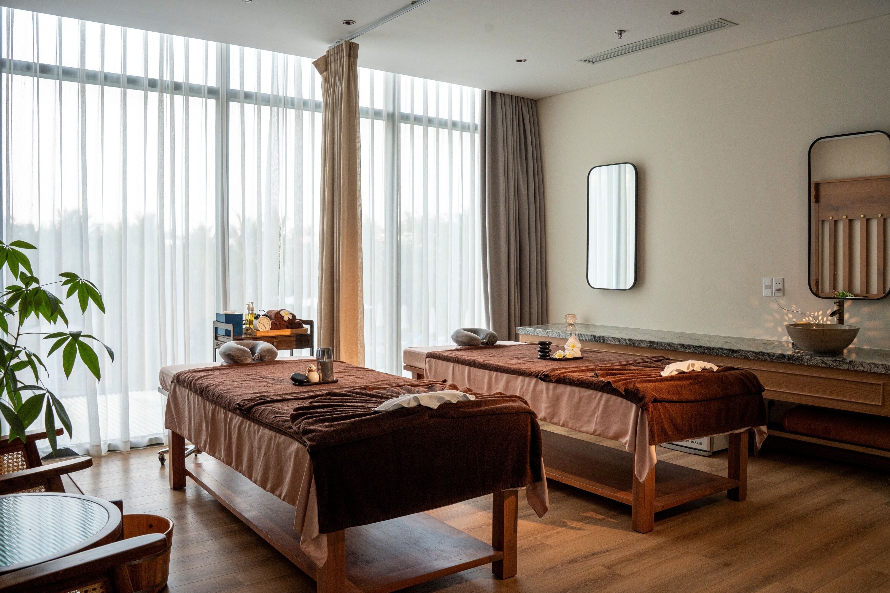 Ein Raum mit zwei Massagetischen, einer Holzbank, einem kleinen Tisch mit einem Korb, einem Spiegel und einem großen Fenster mit Vorhängen.