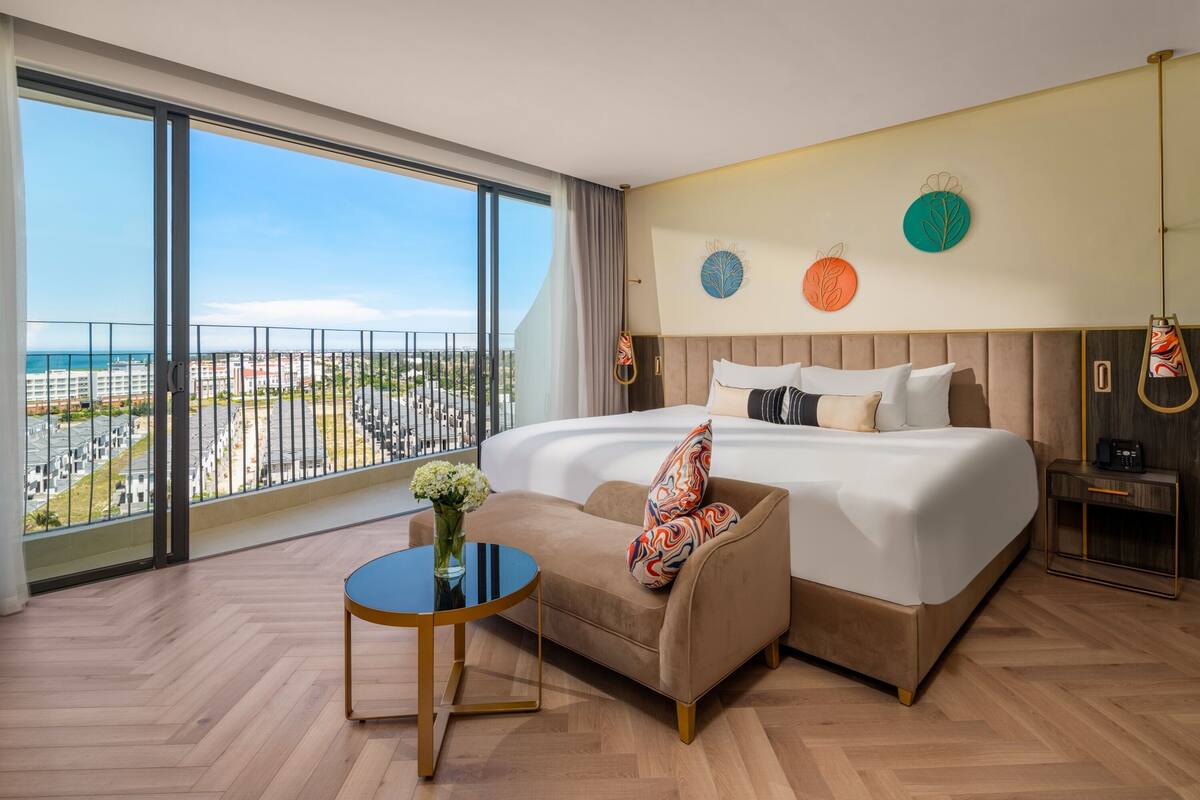 A hotel room with a large bed, a sofa, a small table, and a view of the city through large windows.