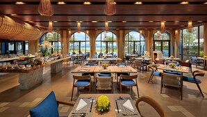 A spacious dining area with multiple tables and chairs, large windows offering a view of the outdoors, and hanging pendant lights.