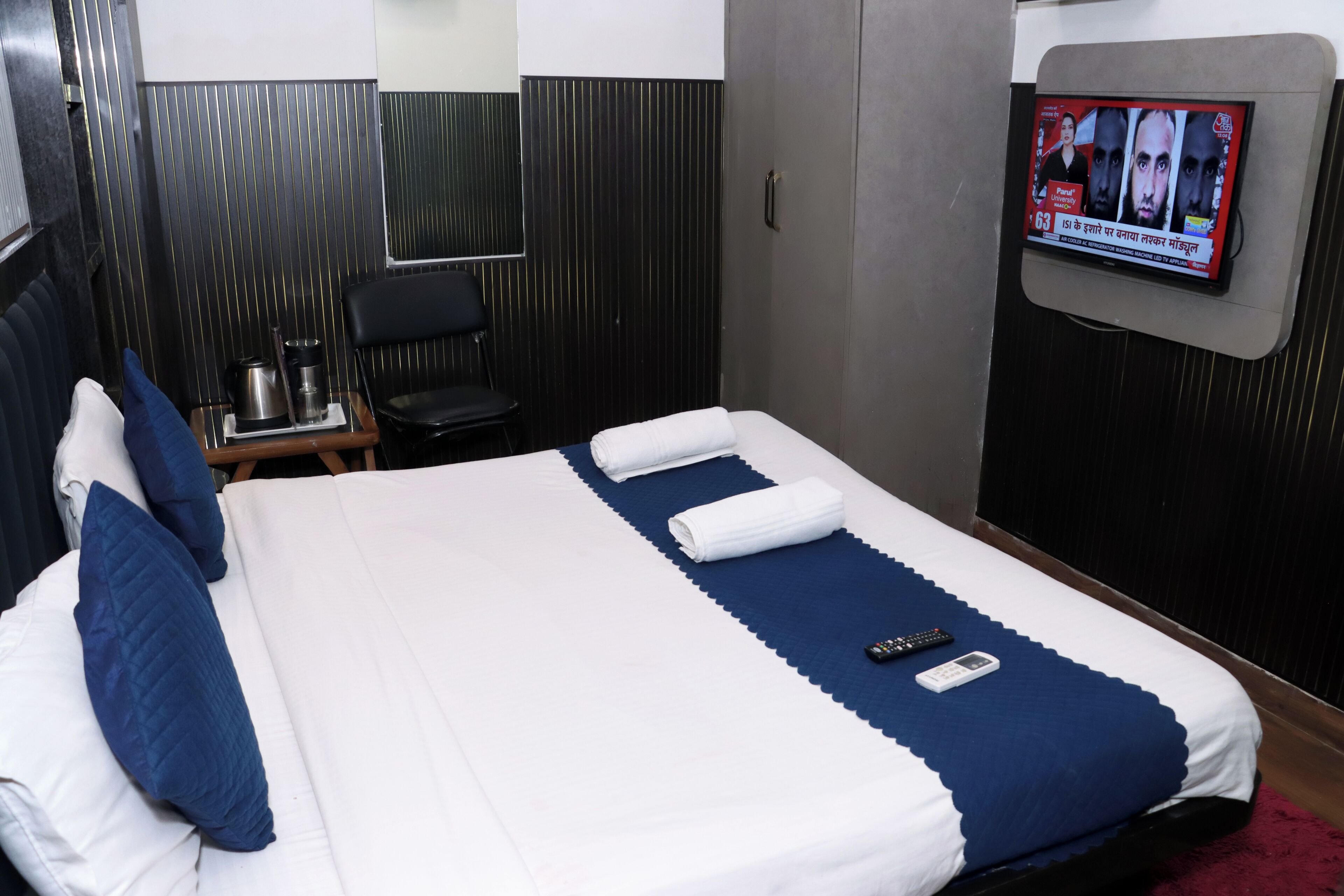 Habitación de hotel con cama, televisor montado en la pared, escritorio con silla y una mesita con hervidor.