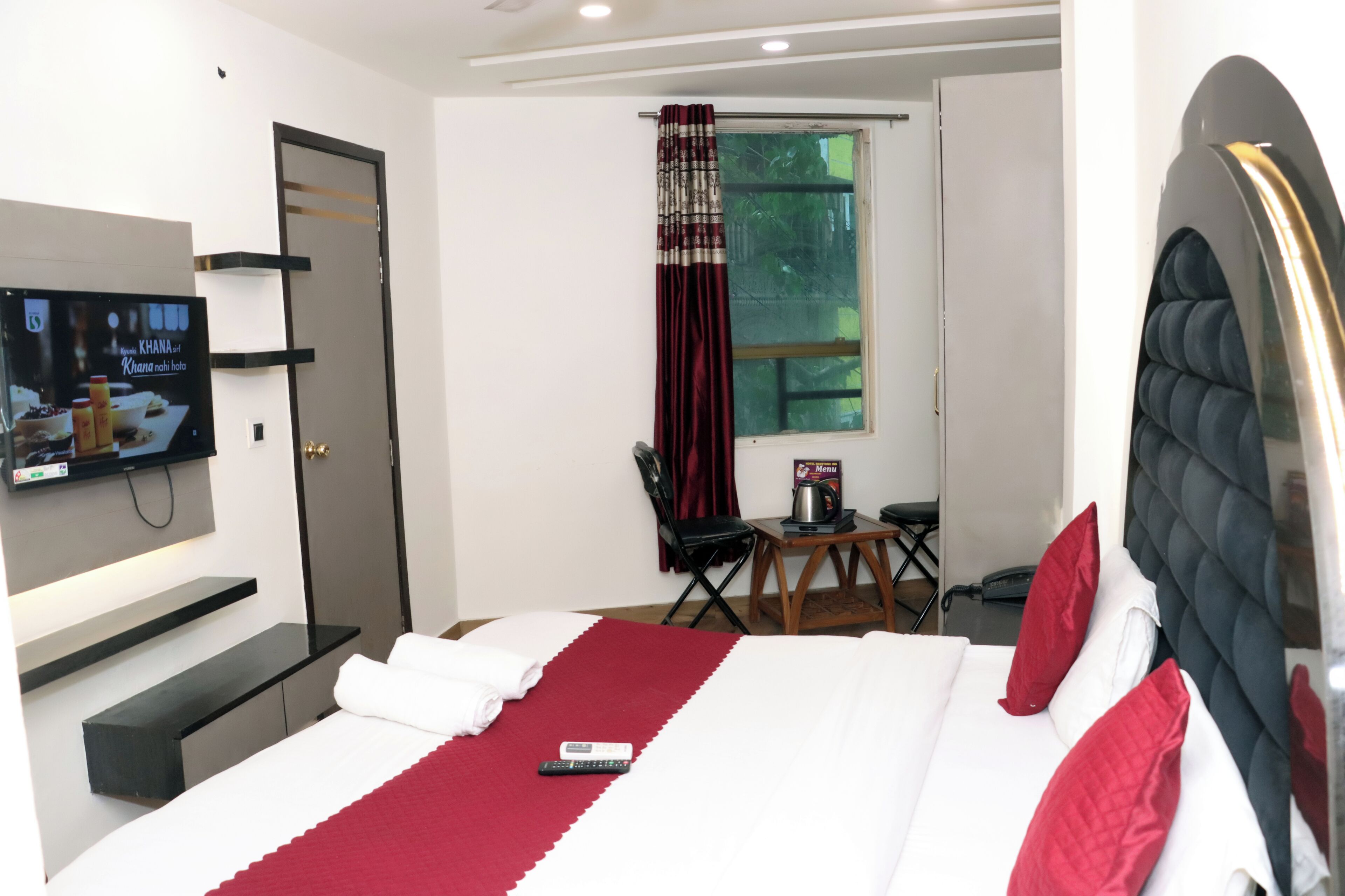 A hotel room with a bed, a TV mounted on the wall, a desk with a chair, and a window with curtains.
