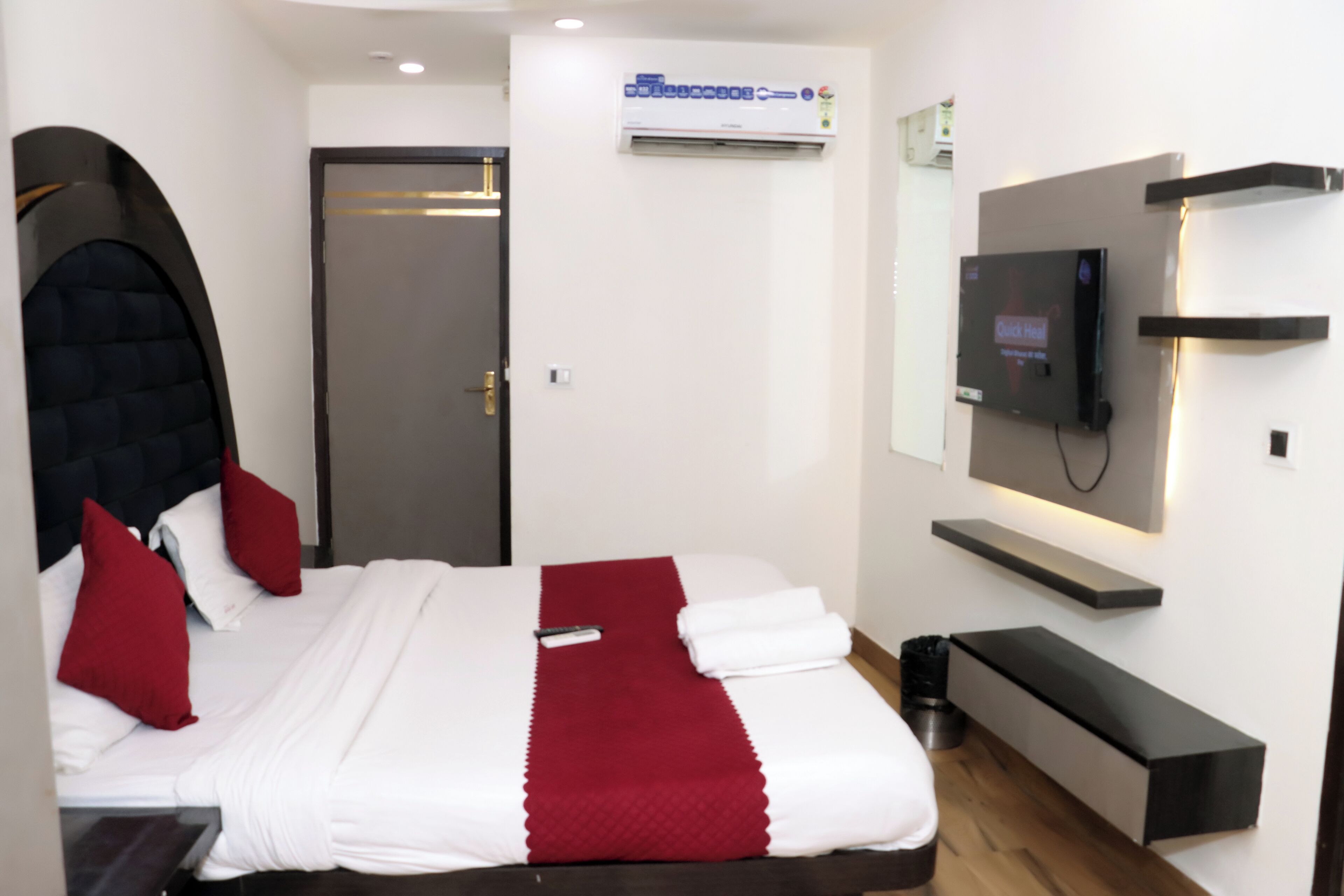 A modern hotel room with a bed, a TV, a wall-mounted shelf, and an air conditioning unit.