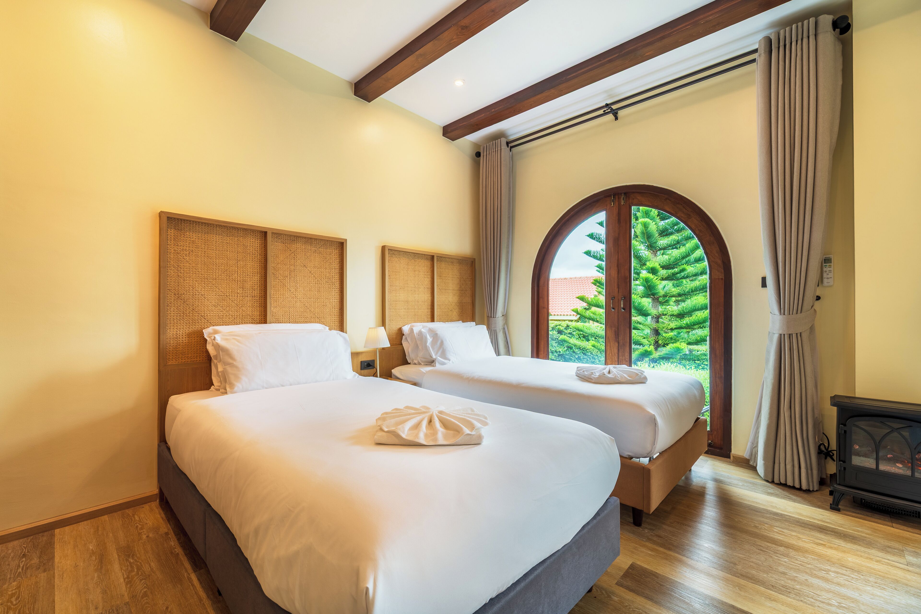 A hotel room with two beds, a wooden ceiling, a large window with curtains, and a fireplace.