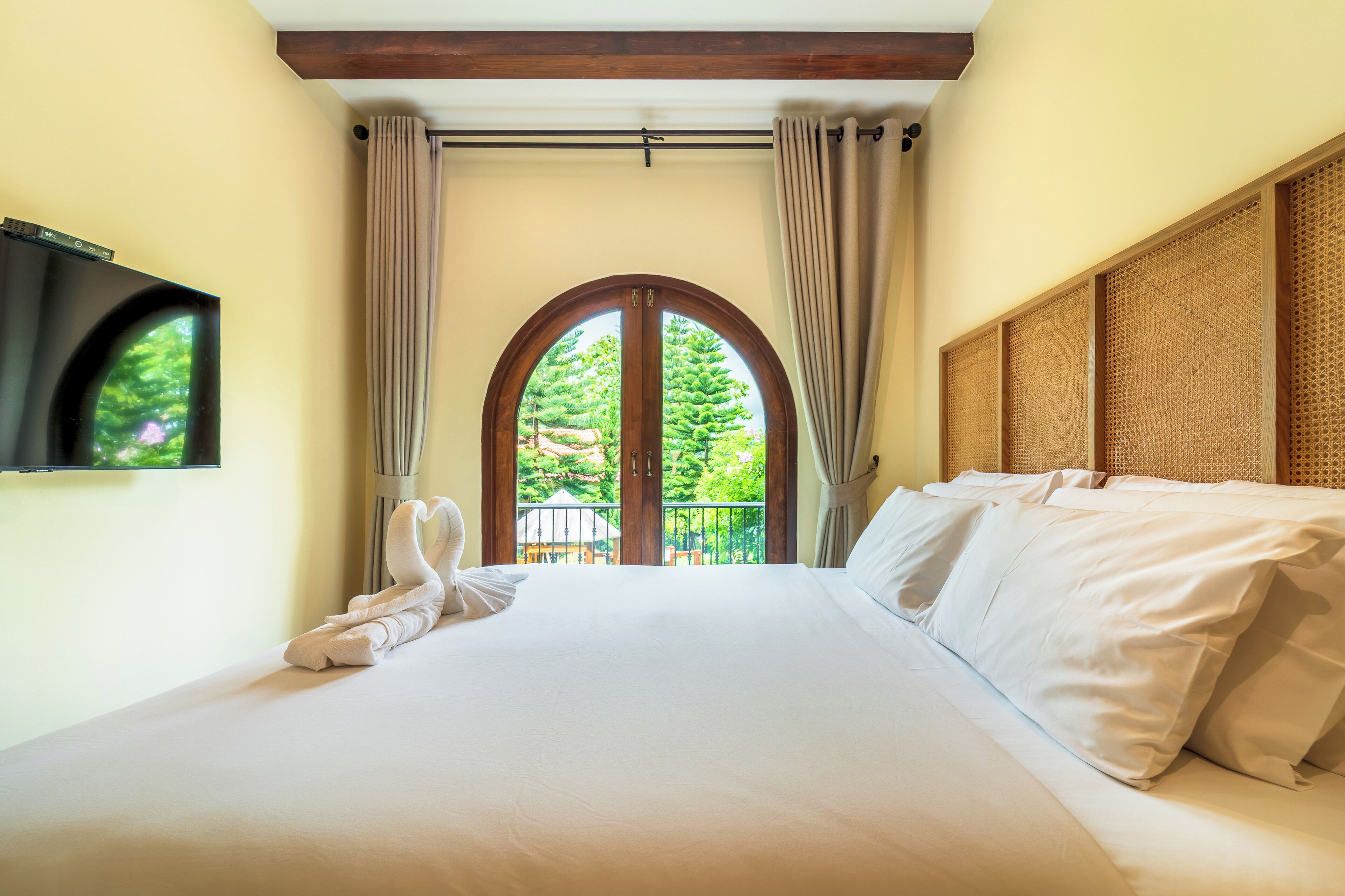 A neatly made bed with white linens, a television mounted on the wall, and a balcony with a view of greenery.