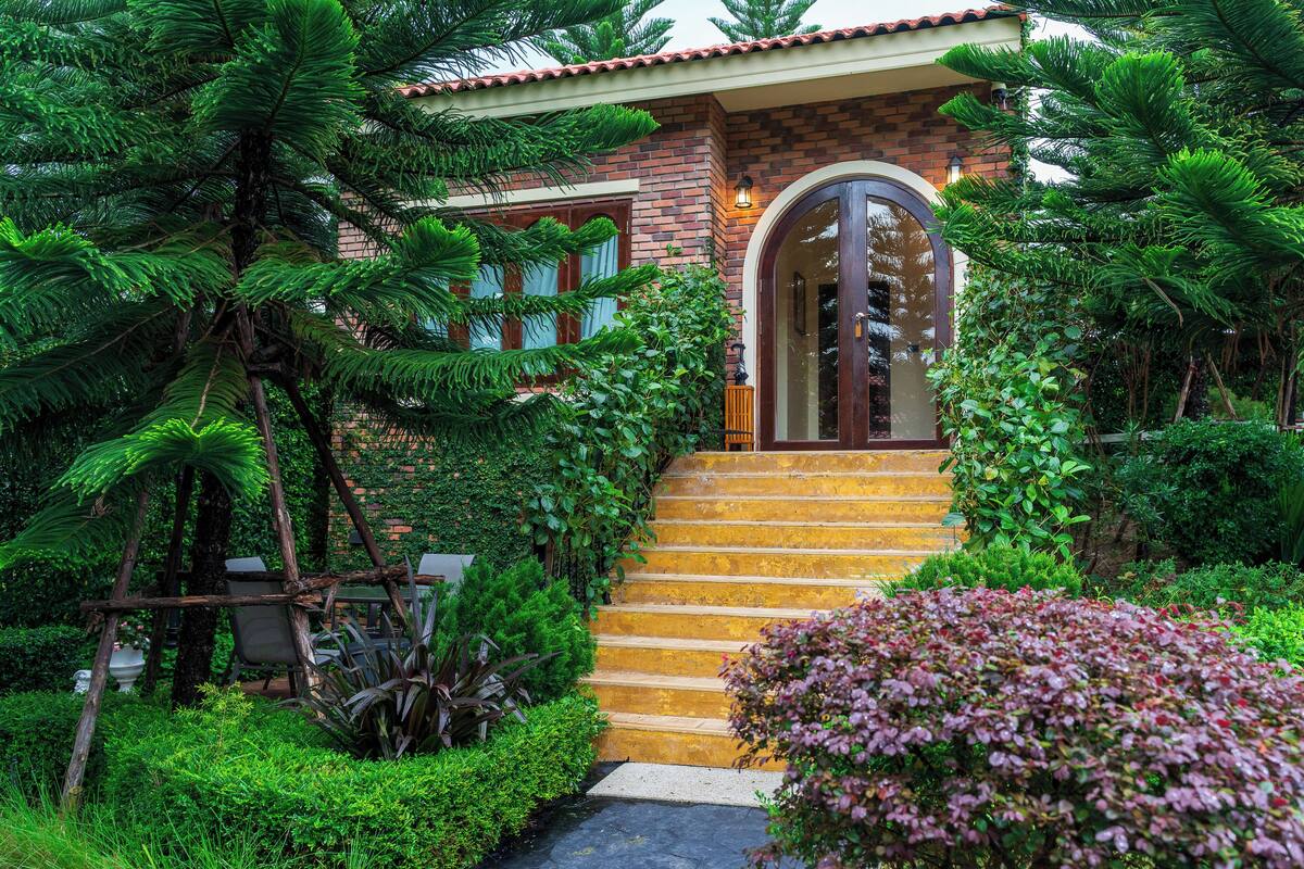 Una casa de ladrillo con una escalera de madera que lleva a una puerta de cristal, rodeada de abundante vegetación.