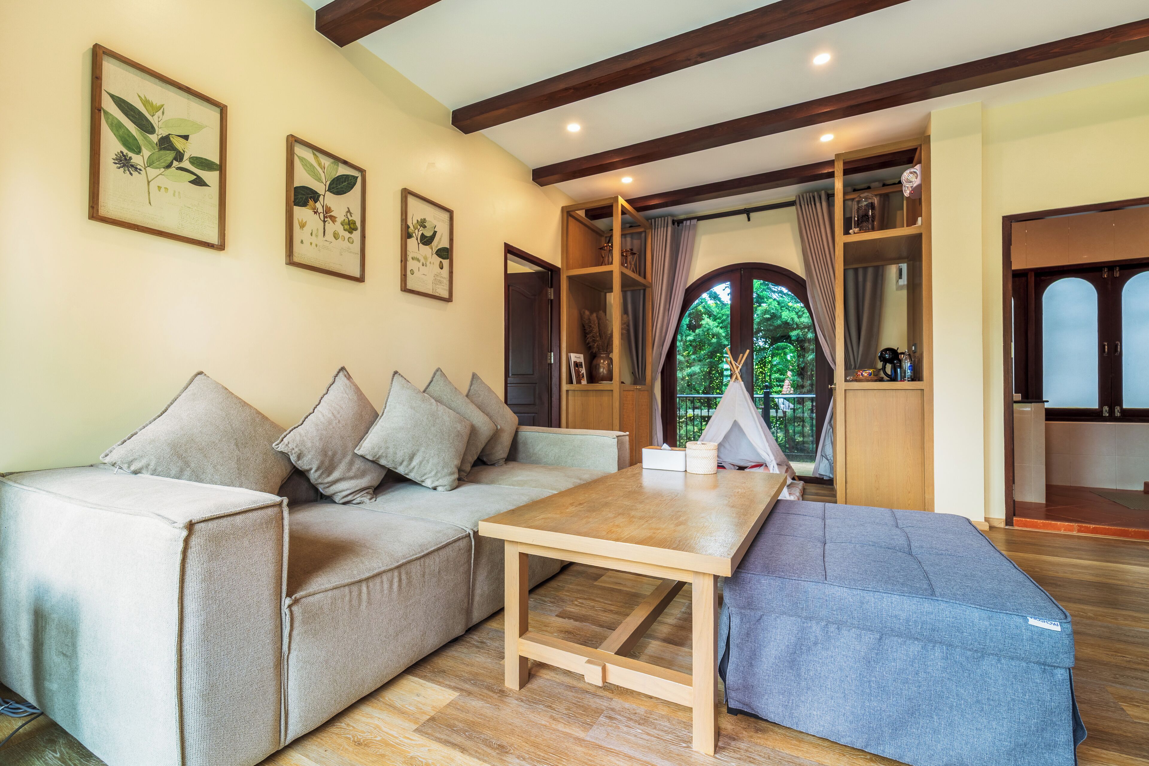 Una sala de estar acogedora con un sofá, una mesa de madera y un banco azul. Hay grabados botánicos enmarcados en la pared y una vista al exterior a través de una ventana.