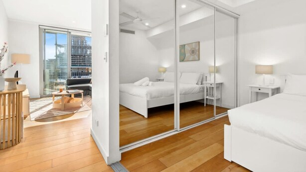 Una habitación de hotel moderna con un gran ventanal corredero que da a un balcón, una cama con ropa blanca, una mesita de noche con lámpara y vistas a la ciudad.