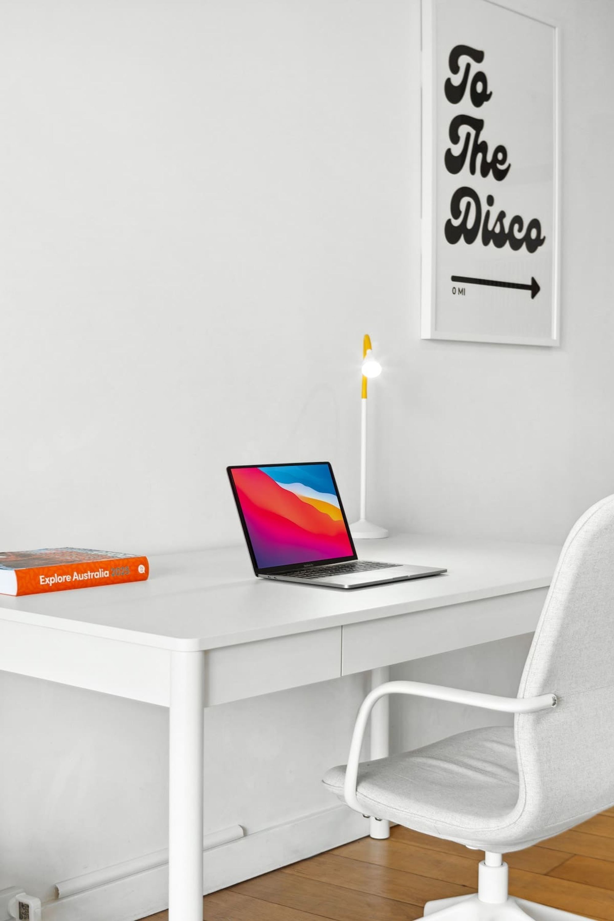 Un escritorio blanco con una computadora portátil, un libro titulado 'Explore Australia 2023', una silla blanca y un letrero en la pared que dice 'To The Disco'.