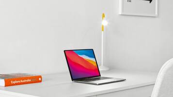 Un escritorio blanco con una computadora portátil, un libro titulado 'Explore Australia 2023', una silla blanca y un letrero en la pared que dice 'To The Disco'.