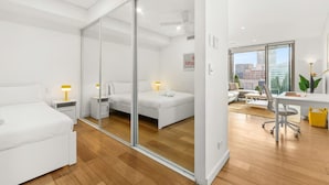 A modern hotel room with a large sliding glass door leading to a balcony, a separate living area with a sofa and a desk, and a large window offering a city view. - Central Luminous 2 Beds With Balcony Aircon (Sydney)