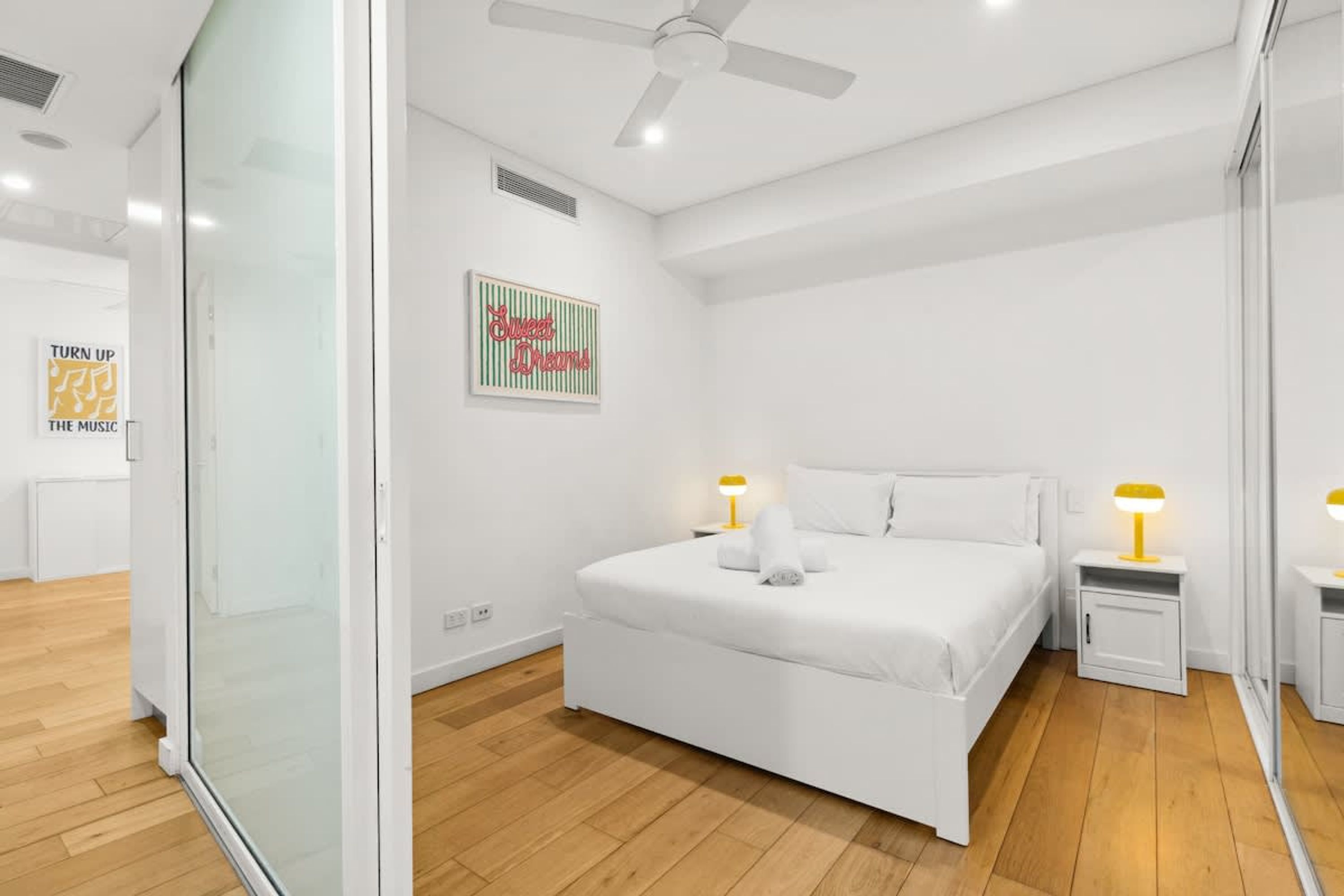 Un dormitori modern amb un llit blanc, dues taules de nit amb làmpades, un ventilador de sostre i una porta de vidre que dona a una altra habitació.