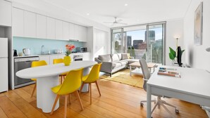 A modern kitchen with white cabinets, a dining area with a white table and yellow chairs, a grey sofa, and a large window with a city view. - Central Luminous 2 Beds With Balcony Aircon (Sydney)
