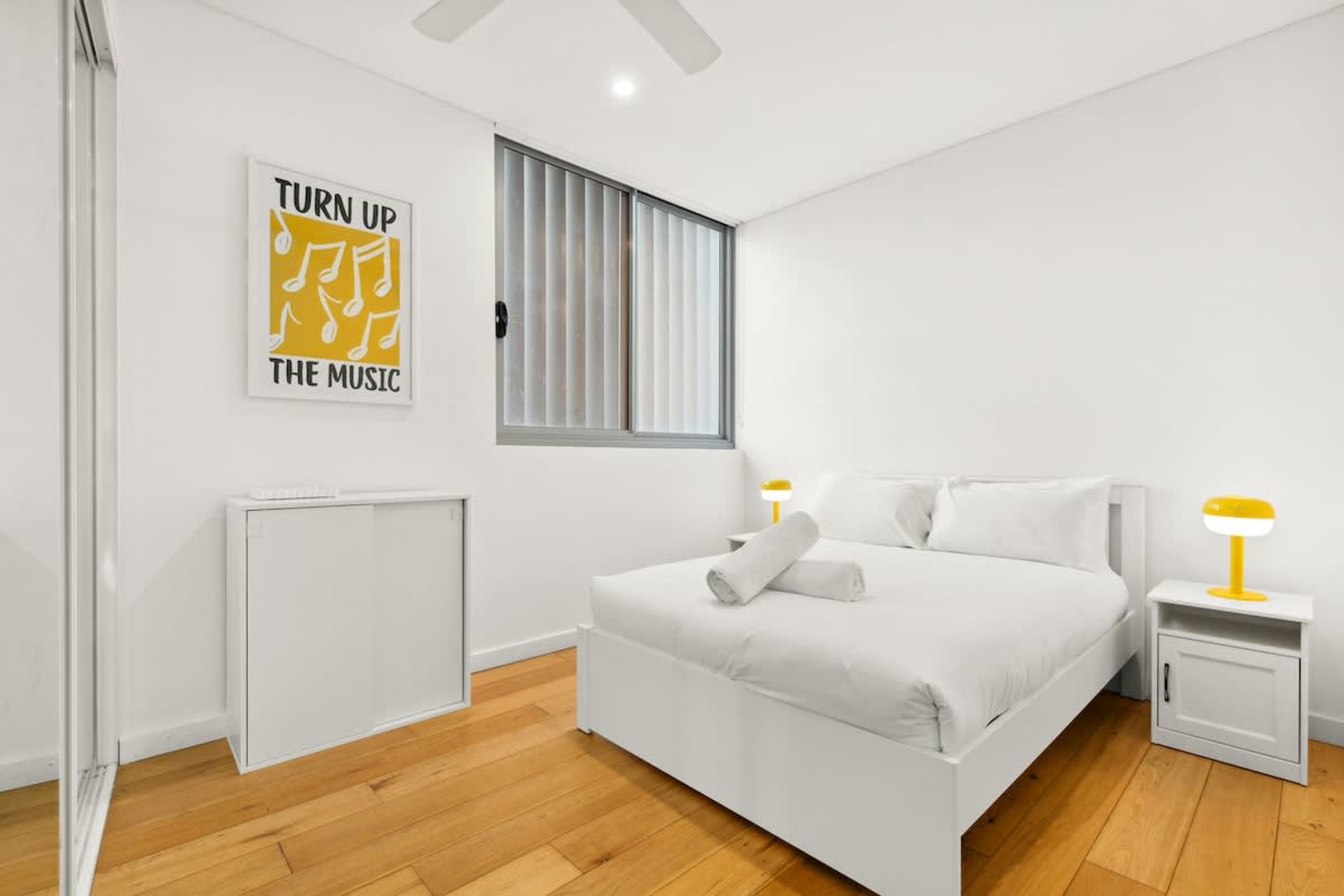 Un dormitorio moderno con una cama blanca, un ventanal con persianas, un radiador y una obra de arte en la pared.