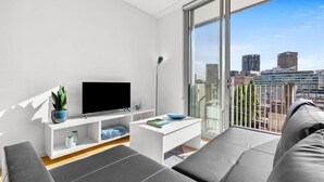 A modern living room with a flat-screen TV, a white entertainment unit, and a large window offering a city view. - Central Art-deco 1bed With Balcony Aircon (Sydney)