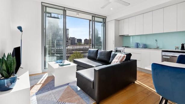 A modern living room with a black sofa, a coffee table, and a TV. There is a large window with a city view. - Central Art-deco 1bed With Balcony Aircon (Sydney)