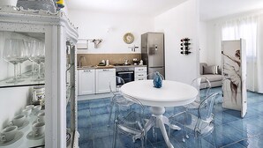 Uma cozinha moderna com armários brancos, uma geladeira de aço inoxidável e um espaço de jantar com mesa branca e cadeiras transparentes.