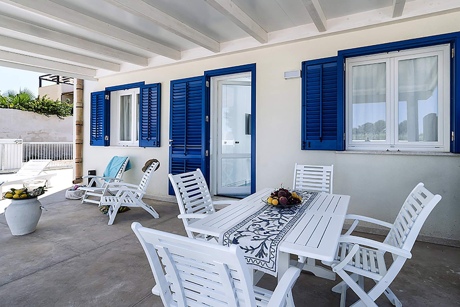 A covered outdoor seating area with white plastic chairs and a table, blue shutters on the windows, and a potted plant on the left.