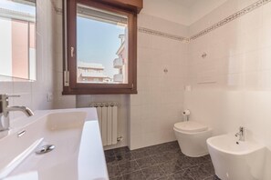Une salle de bain avec un double lavabo, des toilettes et un bidet. Il y a une fenêtre au-dessus du lavabo et les murs sont recouverts de carreaux.