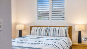 A bedroom with a bed, two bedside lamps, a window with blinds, and a nightstand. - Seadrift 4 on Browning Street (Byron)