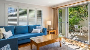 A living room with a blue sofa, a glass coffee table, and a lamp. There is a sliding glass door leading to an outdoor area with chairs and plants. - Seadrift 4 on Browning Street (Byron)