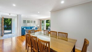 A spacious dining area with a wooden table and chairs, a blue sofa, and a view of greenery through the windows. - Seadrift 4 on Browning Street (Byron)
