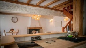 A rustic kitchen with wooden beams, a built-in stove, and a dining area with a round table and chairs. - Les Gîtes Romeline (Sartilly-Baie-Bocage)