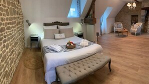 A bedroom with a stone wall, a bed with a headboard, a bench, and a small table with a basket. - Les Gîtes Romeline (Sartilly-Baie-Bocage)