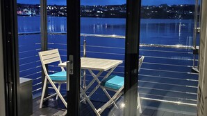 A balcony with a table and chairs, overlooking a waterfront with a bridge and city lights.