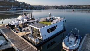 A modern houseboat docked at a marina with a deck, lounge area, and a small boat.