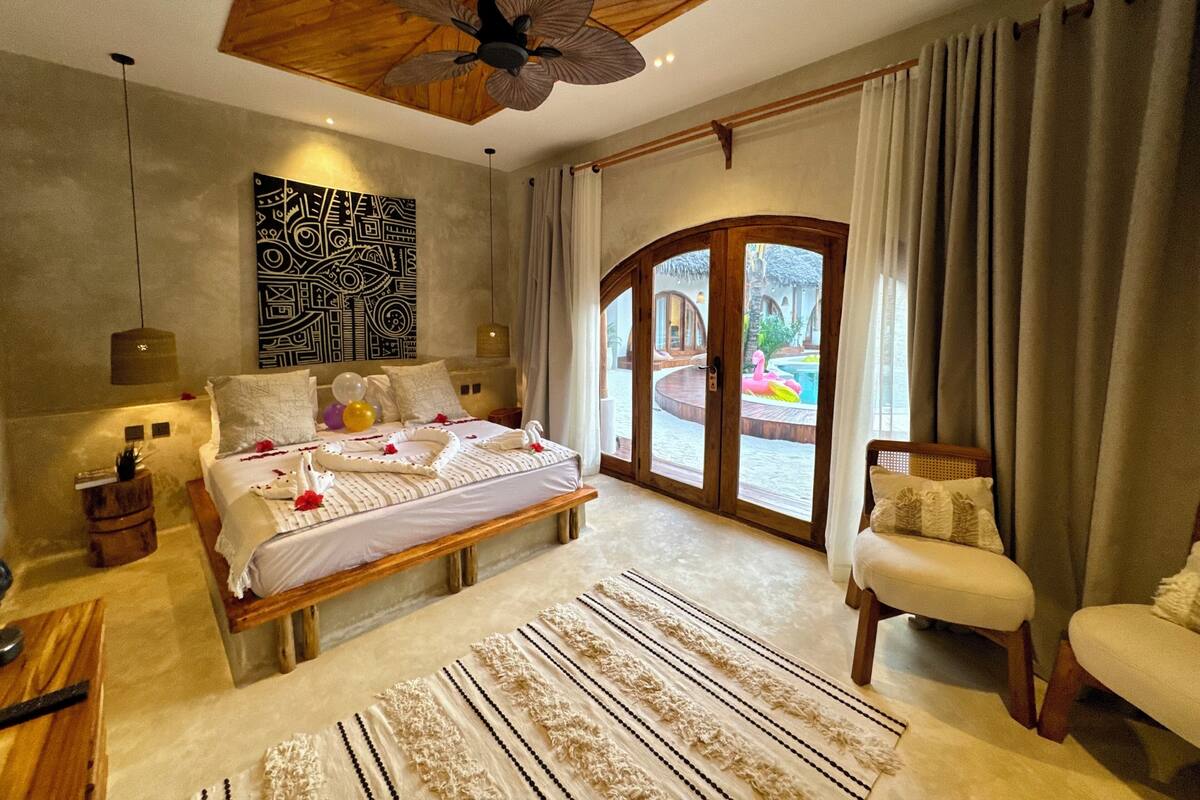 Une chambre spacieuse avec un grand lit, un ventilateur de plafond, une vue sur la piscine et un plafond en bois.
