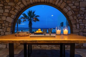Une table dressée avec des citrons, des verres et des bougies, vue à travers une arche en pierre, d’où l’on aperçoit des palmiers et la mer.
