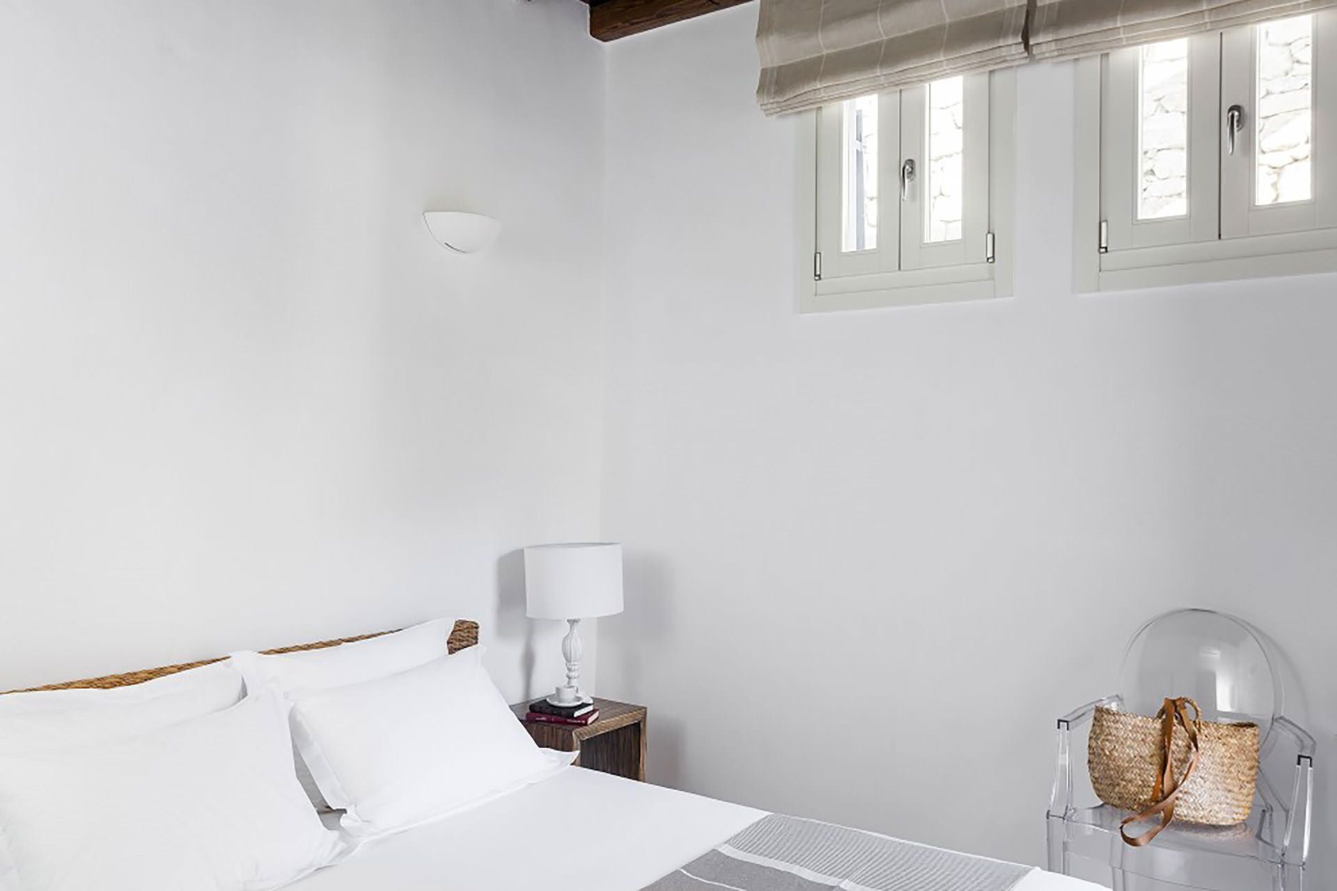 Camera da letto minimalista con un letto, comodino, lampada e una sedia trasparente con una borsa intrecciata.