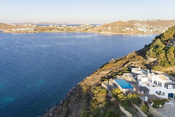 A coastal hotel with a pool, overlooking a large body of water and a hilly landscape.