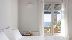 Quarto com cama, mesa de cabeceira, um copo de sumo de laranja e vista para o mar através de uma porta de vidro.