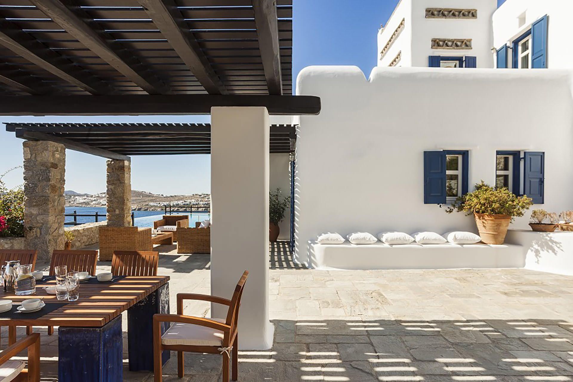 An outdoor seating area with tables and chairs, a white building with blue shutters, and a view of the sea.