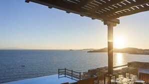 Área de refeições ao ar livre com mesas preparadas para uma refeição, com vista para um corpo d'água e um pôr do sol ao fundo.