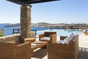 A poolside area with wicker furniture, a stone pillar, and a view of the sea and hills.