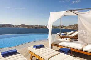 A poolside area with lounge chairs and a canopy, overlooking a body of water and a hilly landscape. - My Rental Homes - Villa Anassa: pool and private dock (possibility of boat rental) (Mykonos)
