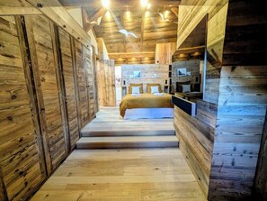 A wooden cabin interior with a bed, built-in shelves, and a staircase leading to the upper level.