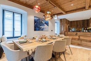 A dining area with a wooden table set for six, surrounded by wicker chairs, with a mountain landscape painting above.