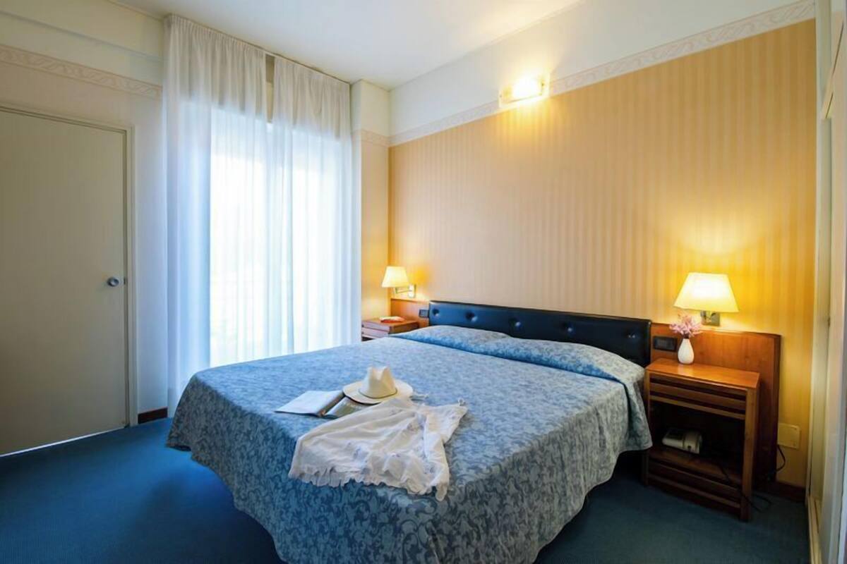 A hotel room with a large bed, two bedside lamps, a nightstand, and a window with curtains.