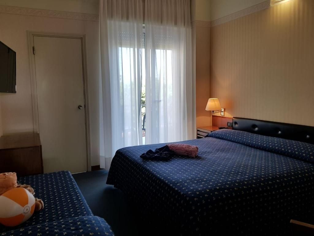 Una habitación de hotel con una cama, una mesita de noche con lámpara, una ventana con cortinas y una puerta.