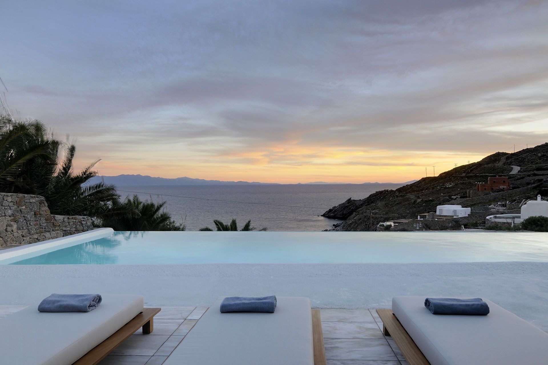 Vista de la piscina con sillas de descanso y un atardecer sobre el mar.
