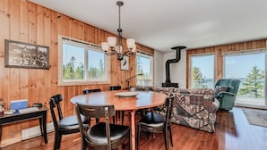 A wooden-paneled room with a round dining table, chairs, a sofa, and a large window offering a view of trees. - Stonehenge  Tobermory - Rustic Waterfront Retreat (Tobermory)