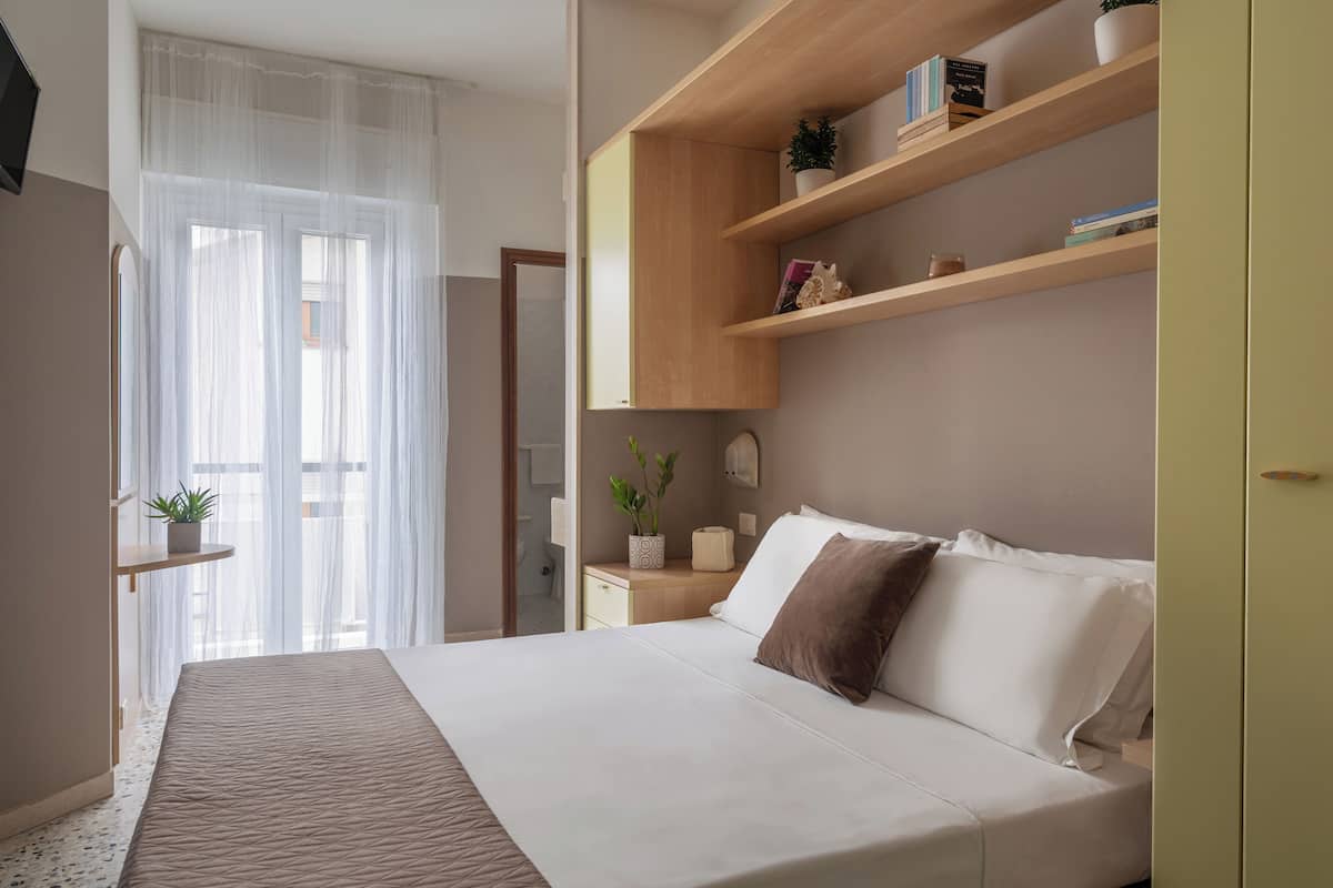 A neatly arranged bedroom with a bed, bedside tables, a shelf with books and a plant, and a window with sheer curtains.