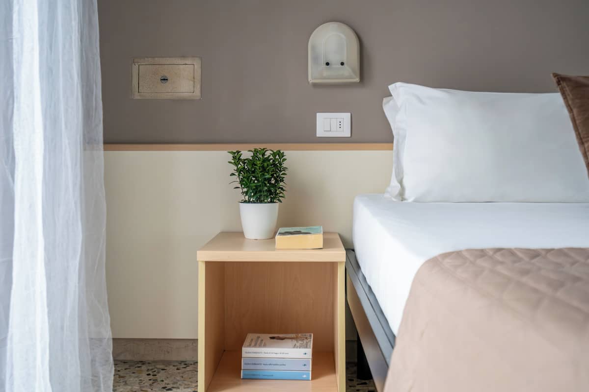A neatly made bed with white bedding, a bedside table with a potted plant, a book, and a wall-mounted light fixture.